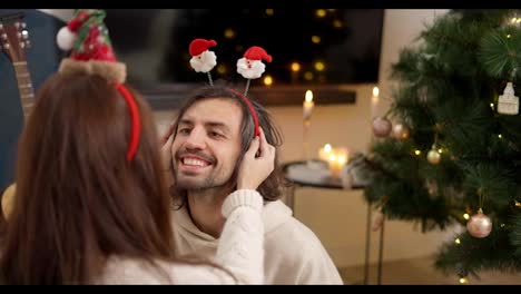 Over-the-shoulder,-a-brunette-girl-puts-on-her-brunette-boyfriend-in-a-white-sweatshirt-a-red-headband-with-Santa-Claus-emoticons-near-a-green-Christmas-tree-in-a-cozy-room-in-the-winter-evening