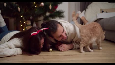 Una-Feliz-Pareja-De-Jóvenes,-Un-Chico-Y-Una-Chica-Vestidos-De-Blanco,-Se-Tumban-En-El-Suelo-Y-Juegan-Con-Su-Gato-Color-Crema-Cerca-Del-árbol-De-Año-Nuevo-Y-Del-Apartamento-Decorado-Al-Estilo-De-Año-Nuevo-En-Una-Tarde-De-Invierno