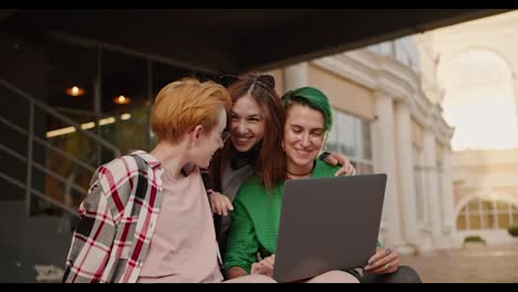 A-trio-of-happy-girls-are-looking-at-something-on-a-gray-laptop-while-sitting-on-a-step-near-a-building-in-the-summer.-A-girl-with-a-short-haircut-with-green-hair-and-a-blonde-girl-with-a-short-haircut-in-a-checkered-shirt,-together-with-her-friend,-a-brunette-girl,-is-looking-at-something-on-the-sc