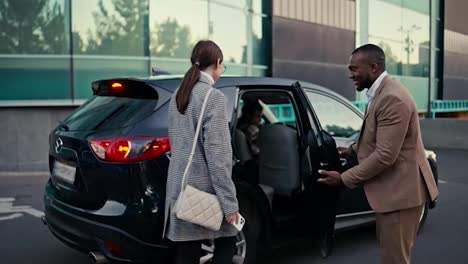 Confident-and-happy-man-Businessman-with-Black-skin-and-a-beard-in-a-brown-suit-invites-a-brunette-female-colleague-to-sit-in-the-passenger-seat-of-a-black-car-and-sits-behind-her-during-their-business-trip-in-the-city