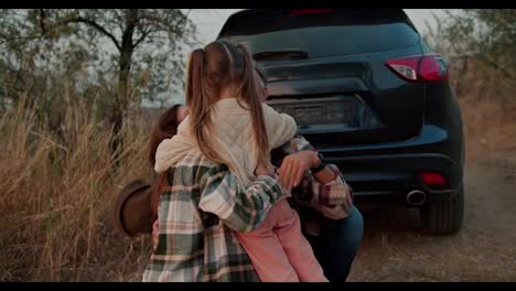 A-little-blonde-girl-hugs-her-parents-near-the-car-in-which-they-arrived-for-a-picnic-outside-the-city-in-the-evening-in-the-summer.-Happy-family-hugging-during-their-picnic-outside-the-city-on-a-summer-evening