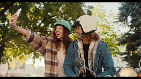 Glückliches-Paar,-Ein-Brünettes-Mädchen-In-Einem-Karierten-Hemd-Macht-Ein-Selfie-Mit-Einem-Telefon-Zusammen-Mit-Ihrem-Freund-Mit-Langen-Lockigen-Haaren-In-Einem-Mopedhelm-Und-Einem-Jeanshemd,-Der-Auf-Einem-Moped-In-Einem-Sonnigen-Sommerpark-Sitzt