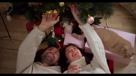 Close-up-shot-of-a-guy-in-a-White-sweater-together-with-his-girlfriend-looking-at-a-Christmas-tree-from-below-near-them-their-cream-colored-cat-pink-gifts-and-a-room-decorated-in-a-New-Year's-atmosphere