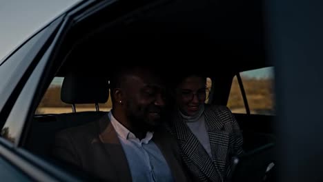 A-happy-man-with-black-skin-in-a-brown-suit-explains-to-a-brunette-girl-in-round-glasses-to-his-colleague-about-his-thoughts-and-ideas-using-a-tablet-during-a-shared-trip-in-a-modern-car