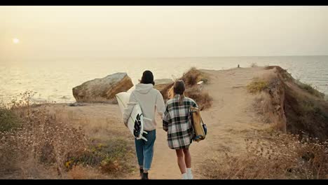 A-happy-brunette-guy-in-a-black-hat-in-a-white-sweatshirt-and-his-blonde-girlfriend-in-a-checkered-shirt-walk-down-the-slope-to-the-sea-and-hold-surfboards-in-their-hands-near-the-sea-in-sunny-weather-in-the-morning-in-autumn