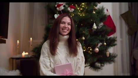 Retrato-De-Una-Niña-Morena-Feliz-Con-Un-Suéter-Blanco-Que-Sostiene-Una-Caja-De-Regalo-Rosa-Cerca-De-Un-árbol-De-Navidad-Decorado-En-Una-Acogedora-Casa-De-Invierno.