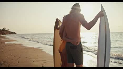 Rear-view-of-a-happy-blond-guy-in-a-hat-with-a-bare-torso-and-in-dark-green-shorts-stands-with-his-blonde-girlfriend-in-an-orange-swimsuit,-they-hold-surfboards-standing-on-a-sandy-beach-and-look-into-the-sea-at-Sunrise-in-the-morning