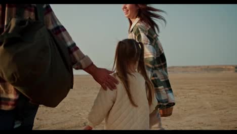 Rear-view-of-a-happy-family,-a-middle-aged-man-in-a-checkered-shirt-walks-along-the-seashore-with-his-wife,-a-brunette-girl,-and-a-little-blonde-daughter-in-a-white-jacket-outside-the-city-during-their-picnic-and-hike-in-the-summer