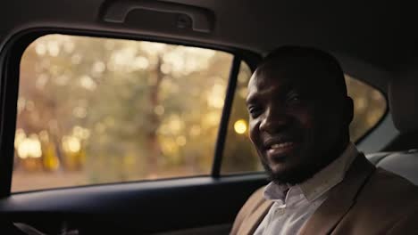 Portrait-of-a-Happy-man-with-Black-skin-and-a-beard-in-a-brown-suit-and-a-white-shirt-who-rides-in-the-passenger-seat-during-his-business-trip-in-a-modern-car-outside-the-city-during-the-sunny-day