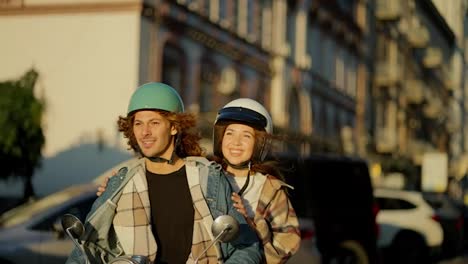 Happy-couple-riding-a-motorcycle,-a-brunette-guy-with-curly-long-hair-in-a-denim-jacket-and-his-brunette-girlfriend-with-long-hair-in-a-checkered-shirt-ride-through-the-summer-city-and-look-at-the-camera