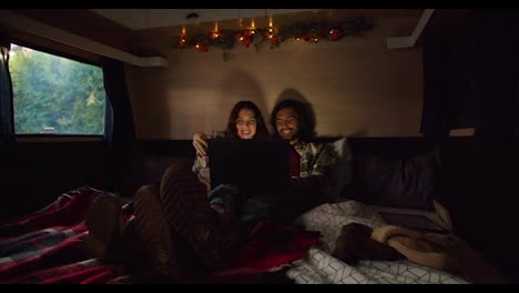 Shooting-in-an-atmospheric-trailer-with-dim-lighting,-a-happy-brunette-couple-in-plaid-shirts-lie-in-their-shoes-on-the-trailer-sofa-and-watch-a-movie-through-a-laptop-Near-their-camp-outside-the-city-in-the-summer