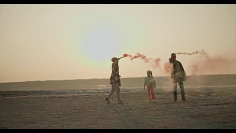 A-happy-brunette-girl-in-a-green-checkered-shirt-together-with-her-husband-and-little-daughter-play-with-fireworks-that-emit-green-and-red-smoke-on-the-shore-of-a-deserted-sea-during-their-vacation-outside-the-city-in-the-summer-evening