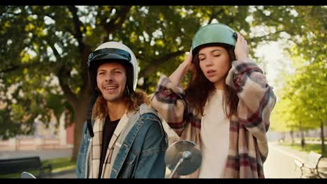 Close-up-shot-of-a-brunette-girl-in-a-plaid-shirt-preening-herself,-looking-into-the-side-window-of-a-moped,-putting-on-a-green-helmet-and-looking-at-her-boyfriend-in-a-denim-shirt-who-smokes-a-cigarette-in-a-White-helmet-while-sitting-on-a-motorcycle-in-a-sunny-city-on-the-street