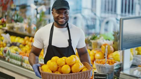 Portrait-of-a-happy-man-with-Black-skin-color-a-supermarket-worker-in-a-white-T-shirt,-black-cap-and-black-apron-holds-in-his-hands-a-large-basket-of-oranges-near-a-counter-with-citrus-fruits-and-berries