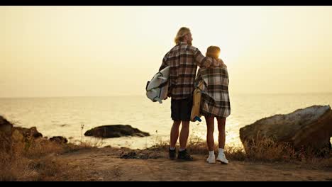 Rear-view-of-a-tall-blond-guy-in-a-plaid-shirt-and-shorts-and-his-blond-girlfriend-in-a-hat-and-plaid-shirt-holding-surfboards-in-their-hands-and-standing-on-the-earthen-seashore-looking-at-the-sunrise-in-the-morning-in-summer