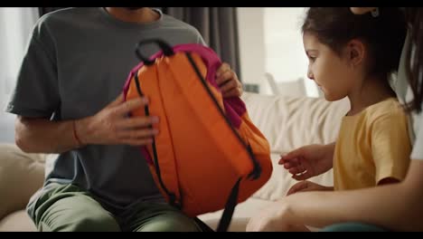 Close-up-shot-of-a-brunette-man-dad-in-a-gray-T-shirt-putting-a-bright-school-backpack-on-his-little-daughter-in-a-yellow-dress.-Happy-mother-of-a-girl-hugs-her,-in-a-modern-apartment