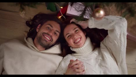 Portrait-of-a-guy-and-a-girl-in-white-sweaters-lying-on-the-floor-near-the-New-Year-tree-holding-hands-smiling-and-looking-at-the-camera-in-a-cozy-room-in-New-Year's-winter