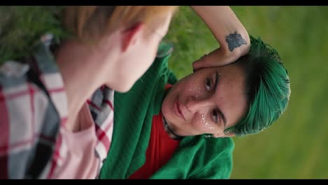 Vertical-video:-a-lesbian-girl-with-green-short-hair-in-a-green-shirt-leans-on-her-arm-and-communicates-with-her-girlfriend-with-a-blonde-with-a-short-haircut-in-glasses-in-a-pink-shirt-during-her-date-in-the-park-lying-on-the-lawn