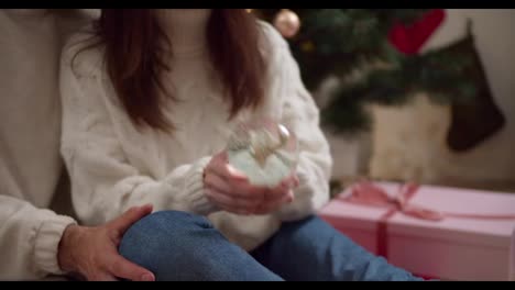 Close-up-shot-of-a-brunette-girl-in-a-White-sweater-shaking-a-New-Year's-glass-sphere-Near-her-boyfriend-in-a-cozy-room-decorated-for-the-New-Year-in-winter