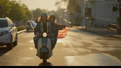 Happy-couple,-a-brunette-guy-with-curly-long-hair-in-a-denim-jacket-and-his-girlfriend-in-a-plaid-shirt-holds-a-large-USA-flag,-which-flutters-beautifully-in-the-wind-while-driving-through-a-summer-city-in-the-morning