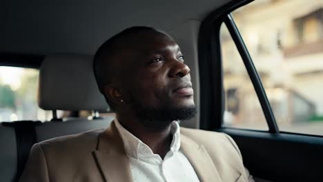 A-man-with-Black-skin-and-a-beard-in-a-brown-suit-rides-in-a-modern-car-and-mysteriously-looks-out-the-window-while-waiting-for-his-arrival-time.-Confident-male-businessman-driving-in-a-car-around-the-city