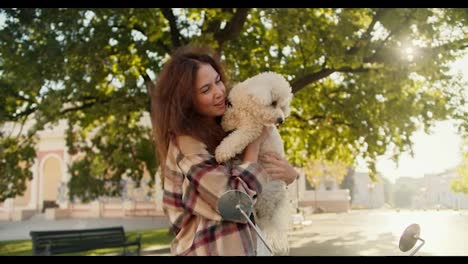 A-happy-brunette-girl-in-a-plaid-shirt-holds-a-curly-small-white-dog-in-her-arms,-smiles-and-looks-at-the-camera-in-a-summer-city-park-near-her-moped