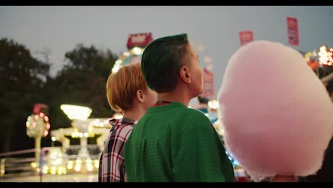 Ein-Mädchen-Mit-Kurzen-Grünen-Haaren-In-Einem-Grünen-Hemd-Und-Ein-Blondes-Mädchen-Mit-Kurzem-Haarschnitt-Halten-Riesige-Rosa-Zuckerwatte-In-Den-Händen-Und-Schauen-Sich-Während-Ihres-Dates-Die-Faszinierend-Leuchtenden-Attraktionen-Im-Vergnügungspark-An