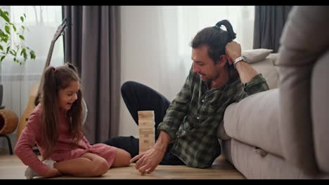 Una-Niña-Morena-Con-Un-Vestido-Rosa-Juega-Con-Su-Padre,-Un-Hombre-Moreno-Con-Una-Camisa-A-Cuadros-Verde,-En-El-Juego-De-Mesa-Jenga-Mientras-Está-Sentado-En-El-Suelo-Apoyado-En-Un-Sofá-Gris-En-Un-Apartamento-Moderno.