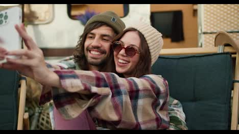 A-happy-brunette-girl-in-round-glasses-and-a-plaid-shirt-together-with-her-brunette-boyfriend-in-a-green-hat-with-stubble-takes-a-selfie-against-the-backdrop-of-a-trailer-and-a-green-chair-during-their-picnic-outside-the-city