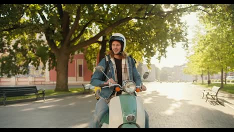 A-happy-courier-guy-with-curly-hair-in-a-denim-jacket-arrived-on-a-moped-to-a-city-park,-looks-around-and-looks-on-his-phone-to-navigate-where-he-should-go-next-to-deliver-his-order-in-a-large-yellow-backpack