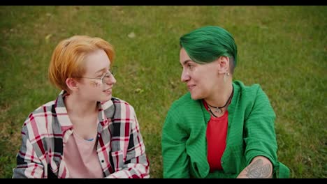 Close-up-shot-of-a-blonde-girl-with-a-short-haircut-with-glasses-in-a-checkered-pink-shirt-and-her-girlfriend-with-a-short-green-haircut-in-a-Green-shirt-and-a-red-T-shirt-are-sitting-on-the-lawn-on-green-grass-and-talking-about-something-during-their-meeting-in-the-park