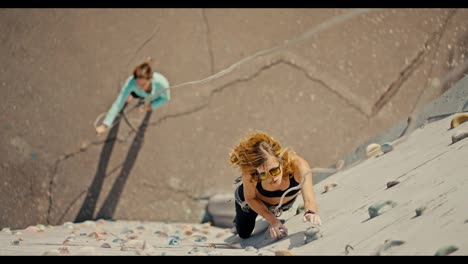 Top-view-of-a-blonde-girl-in-a-black-sports-summer-uniform-and-special-equipment-with-a-belay-climbing-up-a-white-climbing-wall-with-multi-colored-ledges.-And-below-on-the-ground-stands-her-friend-who-belays-the-girl-and-holds-the-rope-to-maintain-the-belay-during-the-climb-and-training-rock-climbin