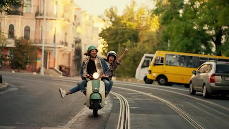 Happy-couple-guy-with-long-curly-hair-in-a-plaid-shirt-and-denim-jacket-rides-with-his-happy-brunette-girlfriend-in-a-plaid-shirt-on-a-moped,-the-girl-puts-her-arms-and-legs-forward-having-fun-and-rejoicing-while-riding-along-a-wide-street-with-her-boyfriend-in-the-summer-city