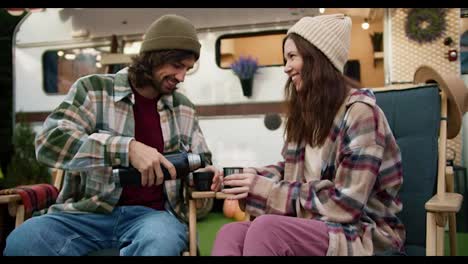 A-happy-brunette-guy-in-a-green-hat-and-a-plaid-shirt-pours-tea-from-a-thermos-to-his-brunette-girlfriend-in-a-hat-and-They-drink-tea-while-sitting-on-green-chairs-near-a-trailer-during-a-picnic-outside-the-city-in-the-summer