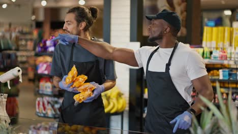 A-man-with-Black-skin-in-a-white-T-shirt-and-a-Black-apron-tells-his-colleague-a-man-with-a-beard-in-a-gray-T-shirt-where-to-put-certain-products-during-their-work-together-in-a-large-supermarket