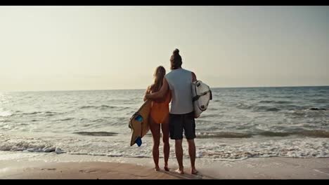 Rear-view-of-a-happy-couple,-a-brunette-man-in-a-white-tank-top-and-his-blonde-girlfriend-in-an-orange-swimsuit-holding-surfboards-standing-on-the-Sandy-Beach-and-looking-towards-the-sea-at-Sunrise-in-the-morning