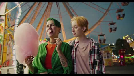 A-girl-with-a-short-green-haircut-in-a-serious-shirt-goes-to-the-half-park-with-her-girlfriend-a-blonde-with-a-short-haircut-in-a-checkered-shirt-and-eats-a-huge-pink-cotton-candy-during-her-date-in-a-bright-and-glowing-amusement-park