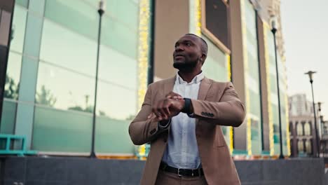 A-confident-man-with-Black-skin-a-short-haircut-and-a-beard-is-persistently-waiting-for-his-colleague-who-is-late-for-a-meeting-in-the-city-and-looks-at-his-watch