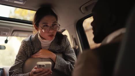 A-confident-brunette-girl-in-round-glasses-in-a-gray-jacket-communicates-with-a-man-with-Black-skin-in-a-brown-jacket-during-her-business-meeting-and-business-trip-in-a-modern-car-interior