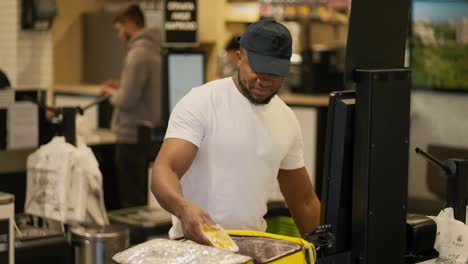 Un-Repartidor-De-Piel-Negra-Con-Una-Camiseta-Blanca-Y-Una-Gorra-Negra-Coloca-Todos-Los-Productos-Necesarios-Después-De-Comprarlos-En-Una-Gran-Bolsa-Amarilla-Sellada-Para-Entregarlos.-Un-Repartidor-Masculino-Compra-Los-Productos-Necesarios-En-La-Tienda.