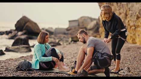 A-blonde-girl-in-a-black-sports-summer-uniform,-a-blonde-girl-with-a-bob-hairstyle-and-a-brunette-guy-in-a-gray-T-shirt-put-on-special-climbing-shoes-and-prepare-to-climb-yellow-rocks-on-a-stony-and-rocky-seashore-in-the-summer