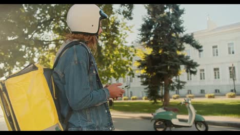 Side-view-of-a-guy-courier-with-curly-hair-in-a-denim-jacket-and-a-white-helmet-for-a-moped-walks-along-with-a-large-yellow-bag-on-his-shoulders,-sits-on-his-green-moped-and-goes-to-deliver-an-order-in-a-sunny-city-park-in-the-summer