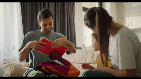 Happy-young-parents-are-getting-their-daughter-ready-for-school-and-packing-a-backpack-for-her,-they-put-different-notebooks-and-textbooks-there-on-a-light-brown-sofa-in-a-modern-room