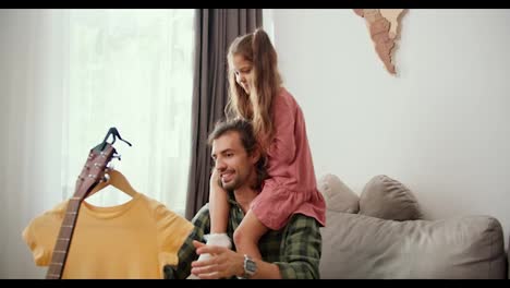 A-little-brunette-girl-in-a-pink-dress-sits-on-the-shoulders-of-her-father,-a-brunette-man-who-is-steaming-his-yellow-dress-in-a-modern-apartment.-A-single-father-communicates-with-his-daughter-while-steaming-a-yellow-dress-at-home-in-a-modern-apartment