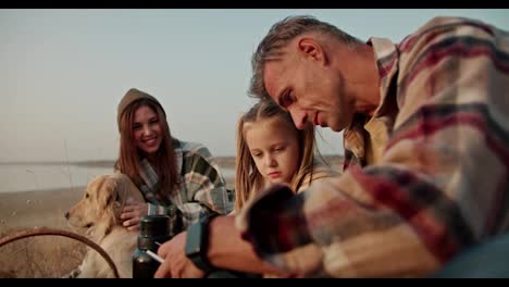 A-happy-brunette-man-with-gray-hair-in-a-checkered-brown-shirt-shows-his-little-daughter-a-blonde-girl-a-book-and-tells-what-it-is-about-during-his-vacation-on-a-picnic-with-his-family,-his-wife-and-a-pet-dog-in-the-summer