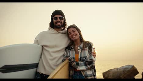 Retrato-De-Una-Pareja-Feliz,-Un-Hombre-Moreno-Con-Gafas-De-Sol-Y-Una-Sudadera-Blanca-Se-Encuentra-Con-Su-Novia-Rubia-Con-Una-Camisa-A-Cuadros-Y-Sostienen-Tablas-De-Surf-Cerca-Del-Mar-Con-El-Telón-De-Fondo-De-Una-Flor-Soleada-Y-Brillante-En-La-Mañana-De-Otoño.