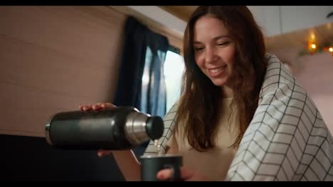 A-happy-brunette-girl-wrapped-in-a-white-blanket-pours-herself-hot-tea-into-a-black-mug-from-a-thermos-while-sitting-in-a-trailer-during-her-picnic-in-the-camp-and-enjoying-the-smell-and-warmth-of-a-hot-drink