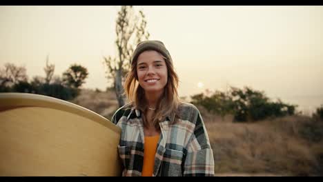 Portrait-of-a-happy-blonde-girl-in-a-hat-in-a-plaid-shirt-who-is-holding-a-wooden-surfboard-in-her-hands-smiling-and-looking-at-the-camera-against-the-backdrop-of-the-sea-and-trees-and-bushes-on-the-slope-of-the-shore