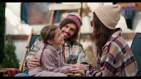 Happy-little-girl-in-a-pink-hoodie-communicates-with-her-mom,-a-brunette-girl-in-a-white-hat-and-plaid-shirt,-and-with-her-dad,-a-man-in-a-green-plaid-shirt,-during-her-vacation-and-picnic-near-a-trailer-outside-the-city-in-summer