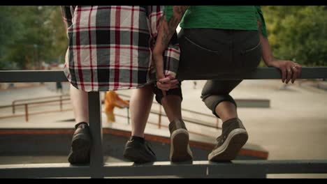 Rear-view-of-a-girl-in-a-plaid-shirt-and-a-girl-in-black-short-jeans-holding-hands-and-sitting-close-to-each-other-during-their-date-at-a-skate-park-in-the-summer.-Two-lesbian-girls-holding-hands